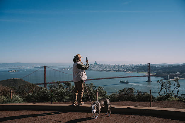 Wide shot of a Filipino man in his 30s stops at a scenic viewpoint and uses his smart phone to take a picture of the Golden Gate Bridge and the San Francisco Bay, while enjoying a relaxing walk with his pet dog on a sunny afternoon.