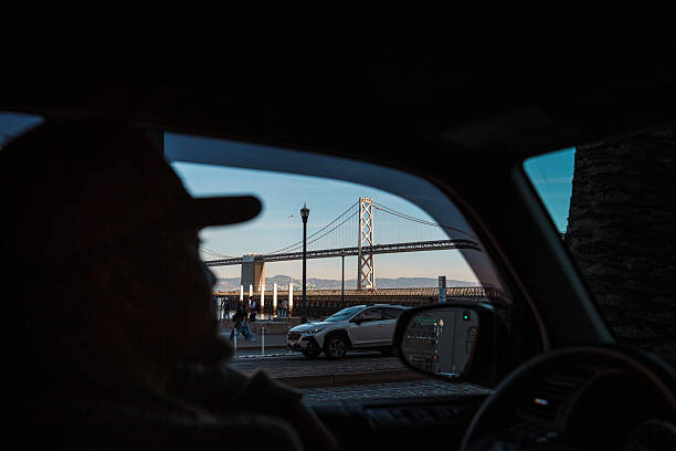 View shot from interior of car past a man in the driver's seat, with selective focus on the Golden Gate Bridge, visible in the distance out the driver's side window on a sunny afternoon in San Francisco, California, USA.