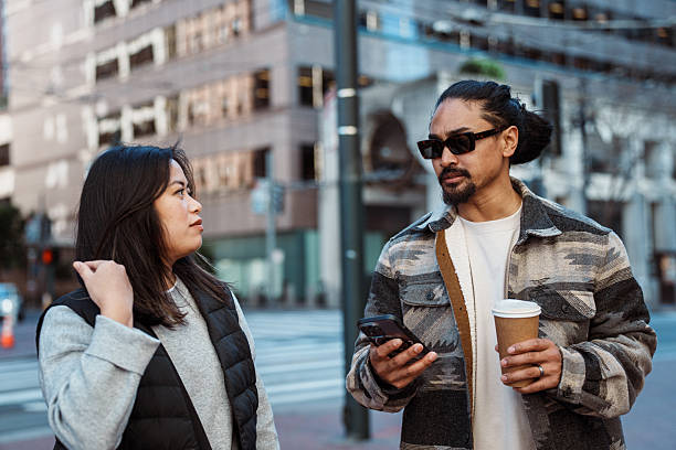 A Filipino man holds a to-go coffee cup and uses his smart phone while talking with his wife as they walk down the street and explore downtown San Francisco, California on a cool afternoon in the winter.