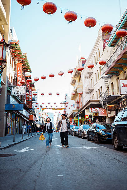 A Filipino couple in their 30s hold hands and walk down a street with red Chinese Lanterns strung overhead while exploring Chinatown in San Francisco, California, USA on a sunny yet cool winter day.