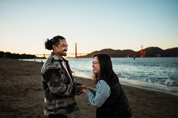 Midrange shot of an affectionate Filipino couple in their 30s laughing together while walking along a sandy beach in San Francisco, California, USA at sunset on a cool winter evening, with a view of the Golden Gate Bridge in the distance.