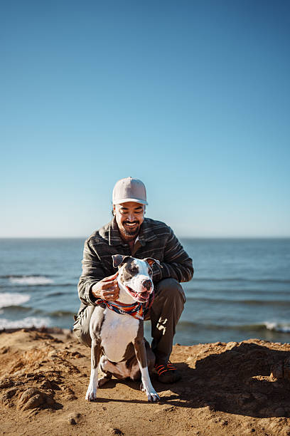 A handsome and fit Filipino man in his 30s squats down and affectionately interacts with his cute and obedient pet dog while enjoying a hike with scenic ocean views near San Francisco, California on a sunny afternoon.