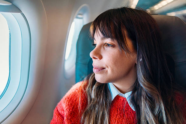 A thoughtful young woman wearing a red sweater gazes out the airplane window during a flight, immersed in contemplation and the journeyâs experience.