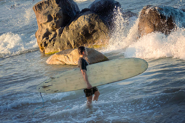 Man holding a surfboard walking through a rocky coastline at the beach