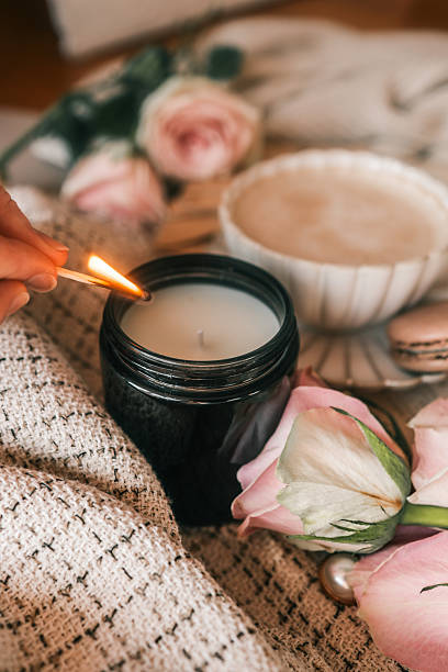 istock A cozy scene with a hand lighting a candle using a matchstick, surrounded by soft beige fabric, a cup of frothy coffee, pink roses, and a macaron. Warm and inviting atmosphere 2206931468