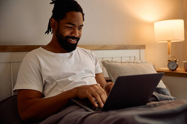 istock Happy black man surfing the net on laptop while relaxing in bed at night. 2206959108