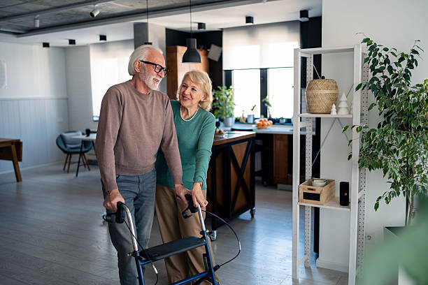 Caring Caucasian Woman Assisting Elderly Husband With Walker Indoors