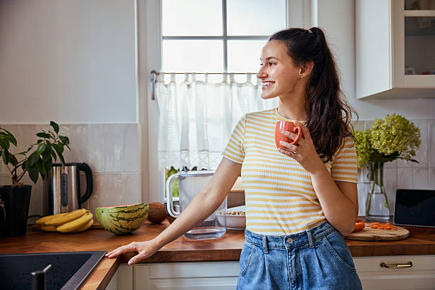 istock Smiling woman in kitchen holding cup looking out the window 2206978336