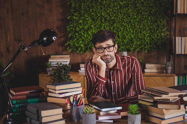 Young man sitting at a desk with books and plants, appearing contemplative, creating a relaxing image suitable for studying, education, or home office themes.