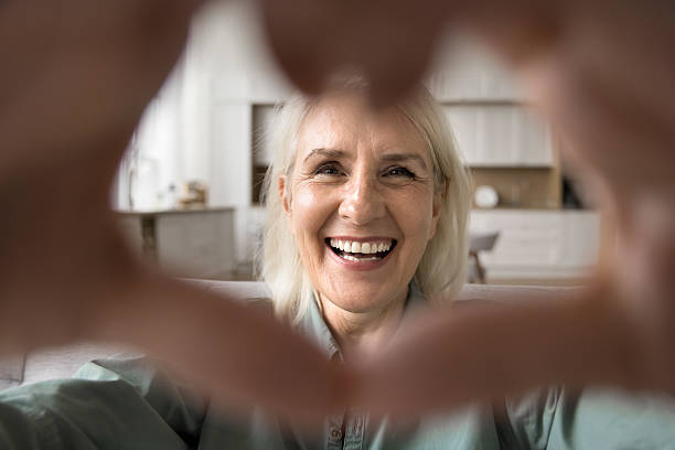 istock Older woman looks at camera forming heart shape with hands 2206988802