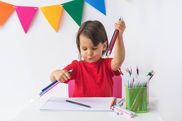 istock Happy child in red t-shirt holding multiple colored pencils while sitting at table with sketchbook, surrounded by art supplies, against background of colorful flags. childhood, creativity, fun 2206988823