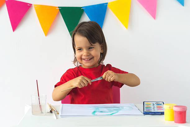 istock Happy child with paintbrush sitting at white table, smiling while creating artwork. Colorful pennant banners in background add festive mood. Concept of childhood, creativity, and artistic expression 2206992232