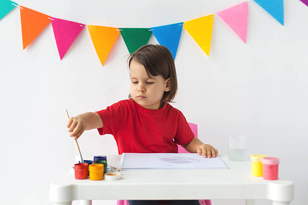 istock A young child in a red t-shirt sits at a white table, reaching for a paintbrush while looking at their drawing on a sketchbook, surrounded by colorful paint jars and water cups 2206992946