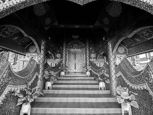 Striking black and white image capturing the intricate details of traditional Thai patterns adorning the entrance to the ubosot (ordination hall) of Wat Phiphat Mongkol in Sukhothai, Thailand. The contrasting tones emphasize the exquisite craftsmanship and artistic heritage evident in every curve and line. The staircase invites viewers to contemplate the spiritual journey within