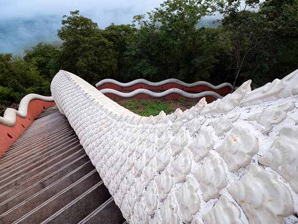 A striking white Naga staircase, resembling the scales of a mythical serpent, winds its way up a mountainside. Lush green forest provides a vibrant backdrop, creating a captivating scene that blends nature and artistic craftsmanship. This image captures the unique beauty of Thai architecture and its connection to mythology