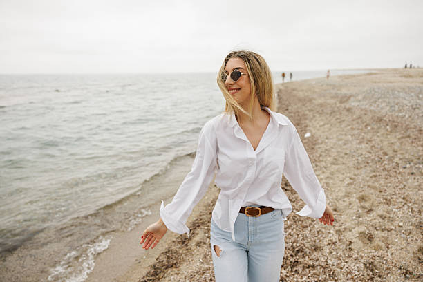 Cheerful young woman in jeans and white shirt enjoying windy day by the sea