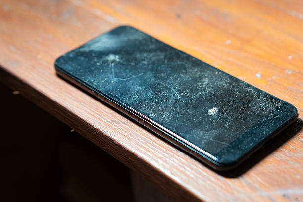 A close-up view of a dusty smart phone lying on a wooden surface. The screen shows visible scratches and smudges, indicating it has not been cleaned for some time.