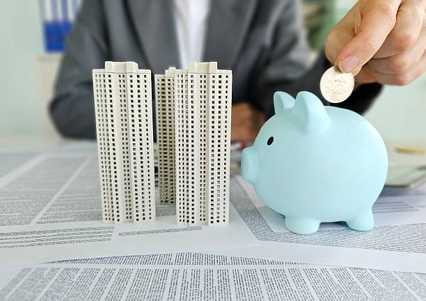 istock A hand drops a coin into a blue piggy bank tall building models sit on a table covered with financial papers concept 2207004966