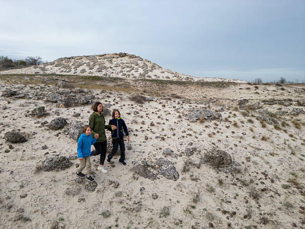 istock Woman walking with two children along a sandy path in a dry, open landscape with scattered rocks and sparse vegetation under a cloudy sky 2207013203