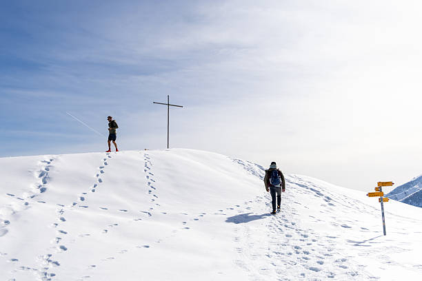Two hikers in snowy landscape near summit cross. Winter trekking in alpine scenery with clear blue sky. Outdoor adventure and exploration in pristine mountain habitat.