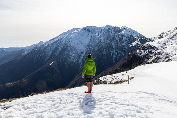 Man in green jacket on snowy peak admiring distant mountains. Winter hiking adventure with majestic alpine landscape. Outdoor enthusiast exploring nature's beauty.