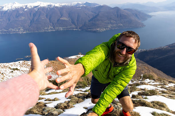 Man Climbing Snowy Mountain Reaching for Help on a Sunny Day Man in green jacket climbing snowy mountain extending hand for assistance. Adventurer enjoying a sunny day with stunning lake view. Outdoor enthusiast in a mountainous landscape. reach destination stock pictures, royalty-free photos & images