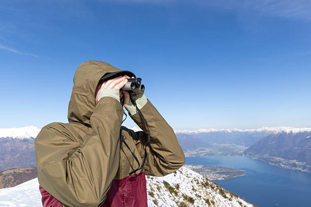 Individual in hooded jacket gazes through binoculars over snowy mountain ridge. Hiker explores scenic winter terrain. Outdoor enthusiast enjoying breathtaking mountainous vista.
