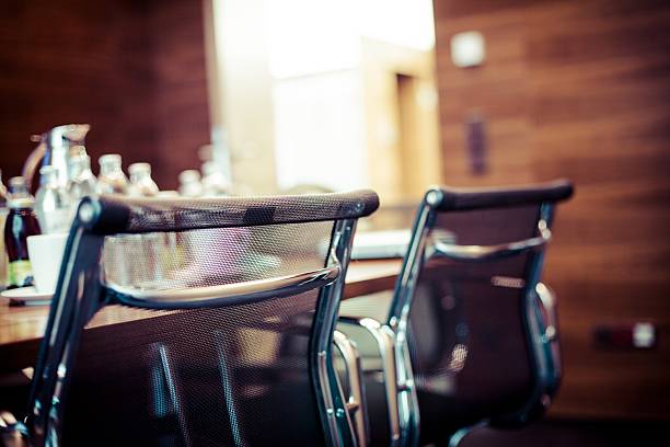Defocused profile view through the arms of multiple gray, modern, leather chairs pushed into a brown, wooden conference table in a brightly lit white room.