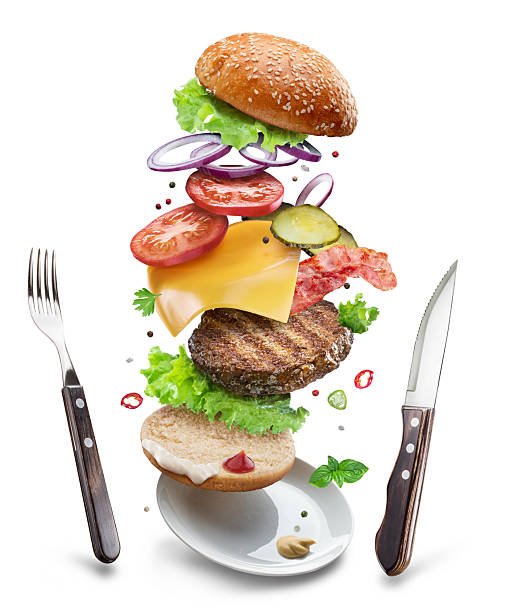 Cheeseburger ingredients with beef patty, vegetables and cheese floating in the air above a plate and cutlery next to it on a white background. File contains clipping paths.