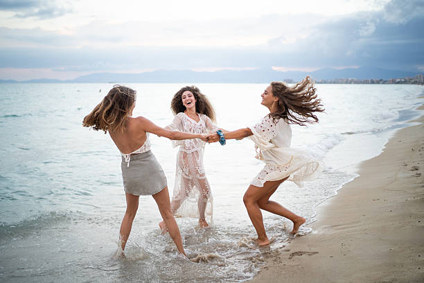 Three carefree young women are holding hands and joyfully spinning around in the shallow water of a beautiful beach, enjoying their friendship and the summer atmosphere