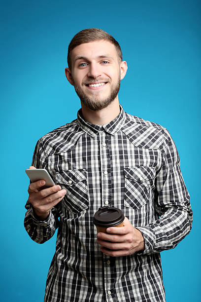istock Young man with smartphone and cup of coffee on blue background 2207032892