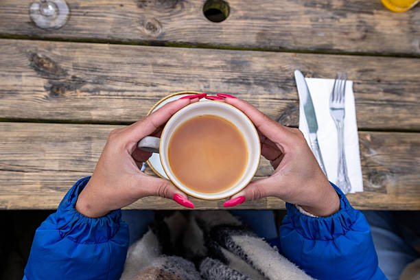Point of view direct above shot of a woman holding a cup of tea in Newton-by-the-Sea, Northumberland. She is sitting outdoors with her warm clothing on, meeting with her friends at a restaurant. Videos are available similar to this scenario.