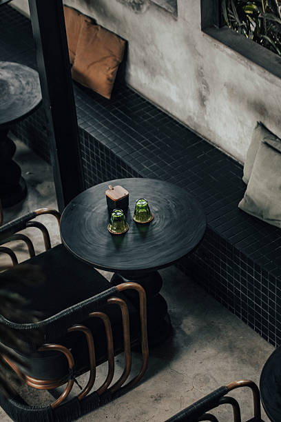 A modern cafe interior featuring a black round table adorned with two small green glass holders. The space includes a tiled bench with cushions and stylish rattan chairs, creating a warm, inviting atmosphere that blends industrial and contemporary design elements.