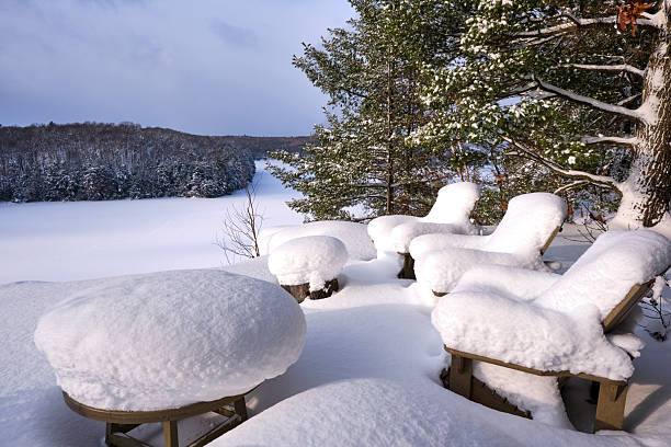 A beautiful winter scene with three muskoka chairs and a table covered in snow overlooking a  Lake in Muskoka.