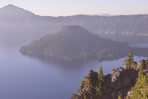 A scenic sunrise view of Wizard Island, a volcanic cinder cone rising from the center of Crater Lake’s vast caldera in Oregon, USA. Soft morning haze veils the surrounding crater rim while foreground cliffs and evergreen trees add striking depth and detail. Formed by the collapse of Mount Mazama over 7,000 years ago, Crater Lake is the deepest lake in the United States and is renowned for its clear blue water and dramatic volcanic geology. This peaceful, layered landscape captures the park’s natural wonder and ancient origins, perfect for themes of geology, national parks, and serene wilderness.
