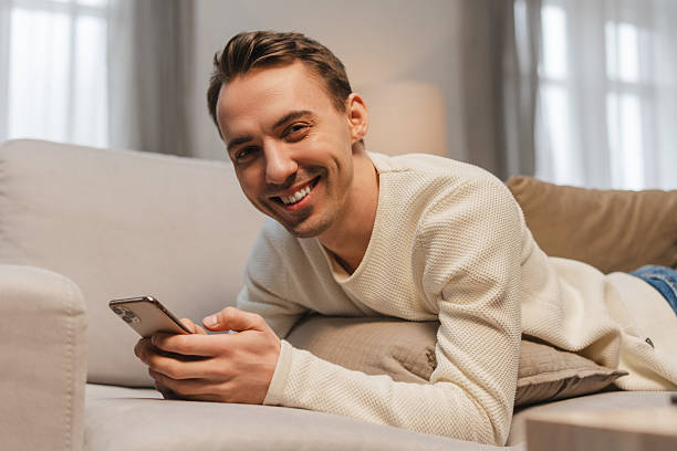 istock Handsome, happy man holding mobile phone, using mobile app, lying on comfortable sofa at home 2207060101