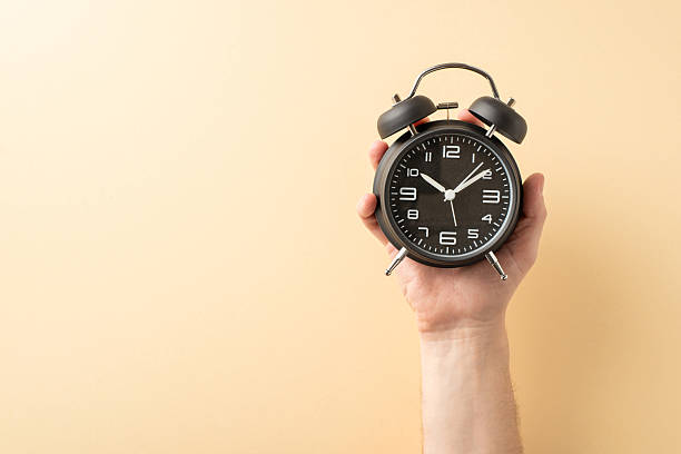 istock Image showcasing a hand holding a black analog alarm clock with a pastel background 2207067783