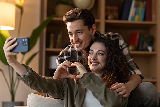 istock Young couple making heart shape with hands while taking a selfie with smartphone at home 2207068713