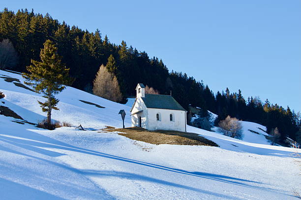 A small white chapel stands alone on a snowy hillside at the edge of a forest, bathed in winter sunlight. Surrounded by stillness and alpine beauty, it evokes solitude and peaceful reflection.