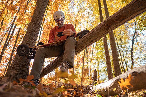 Man with helmet & sunglasses at sunset uses phone sitting on mountain skateboard among vivid autumn forest trees. Wellbeing, technology & sustainable lifestyle.
