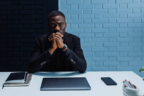 Focused african american entrepreneur reflecting on business strategy while sitting at desk with laptop, smart phone and notebooks in contemporary workspace