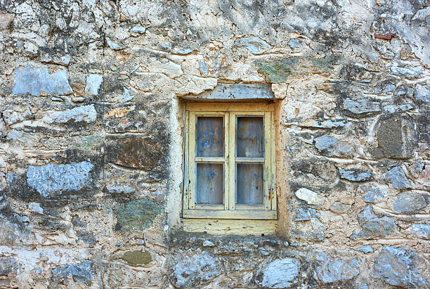 Embedded in a weather-worn stone wall, this rustic wooden window with faded paint exudes timeless allure. The intricate stone patterns tell stories of its past, a perfect motif of rural simplicity.