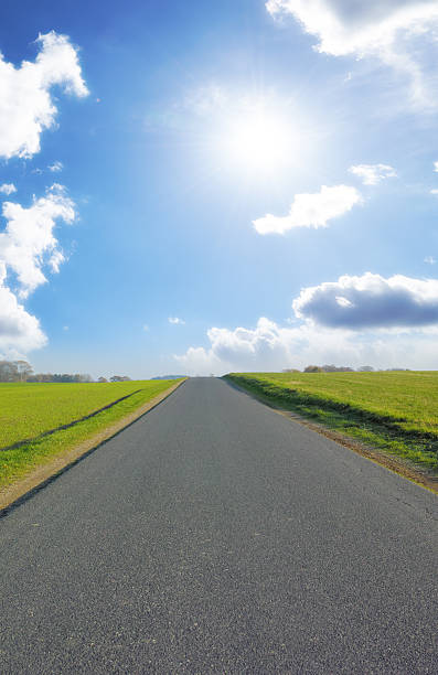 A rural asphalt road stretches into the horizon under a vast and vibrant clear sky dotted with soft, glowing clouds. Lush green fields flank both sides, creating a serene and peaceful scene.