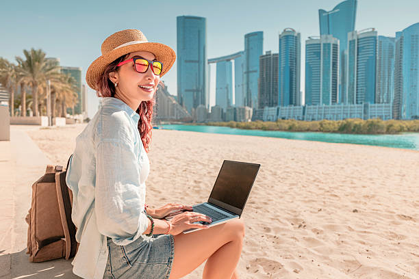 istock Young woman using laptop while sitting on sandy beach with Abu Dhabi skyline in background 2207090955