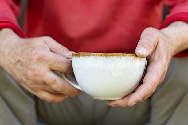 istock A close-up of rough, weathered hands holding a rustic ceramic cup filled with hot tea. The person is wearing a red jacket, and the warm, earthy tones create a cozy and authentic atmosphere. 2207123581