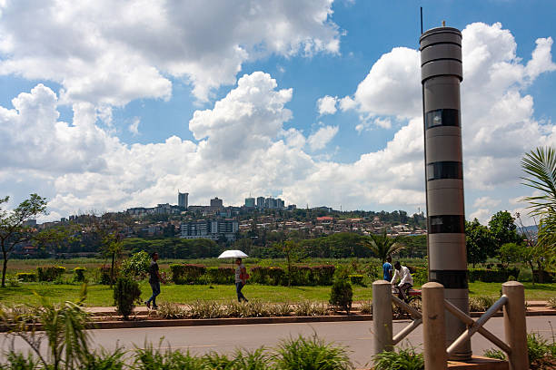 A scenic view of Kigali, Rwanda, showcasing the city's skyline under a partly cloudy sky. In the foreground, a modern traffic surveillance tower stands tall alongside a well-maintained pedestrian walkway, where people are walking, one holding an umbrella. A cyclist is also seen passing by, adding to the city's dynamic atmosphere. The lush greenery and organized urban landscape reflect Kigali's reputation as one of Africa's cleanest and most developed cities.