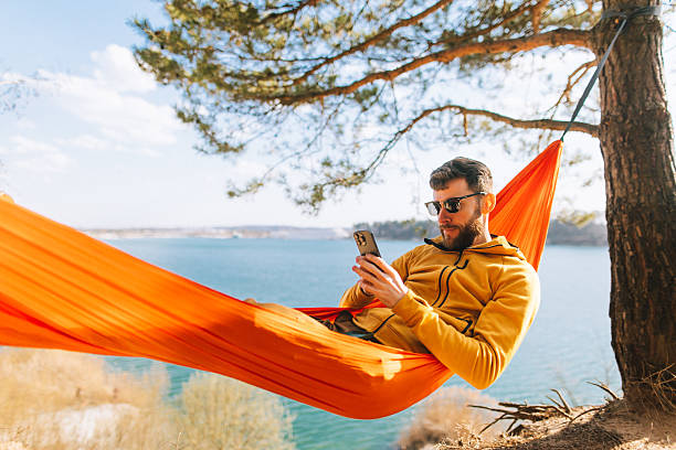 istock Handsome Man Scrolling Smartphone in Lakeside Hammock on Sunny Spring Day 2207131971