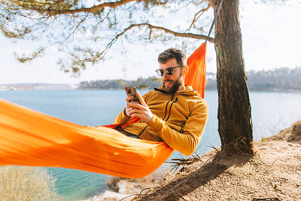 Handsome Man Relaxing in Lakeside Hammock with Smartphone on Sunny Spring Day