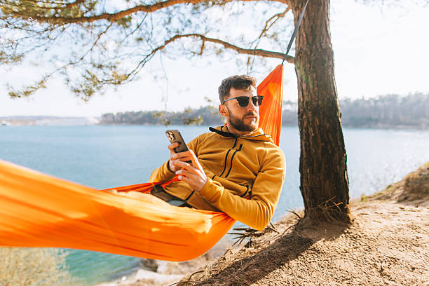 istock Handsome Man Relaxing in Lakeside Hammock with Smartphone on Sunny Spring Day 2207131994