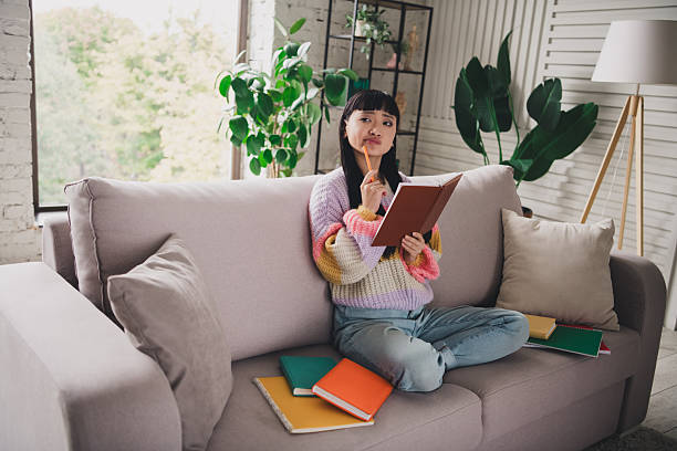 istock Young Asian Woman Reflecting While Writing in a Notebook on a Comfortable Sofa at Home Surrounded by Book 2207139230